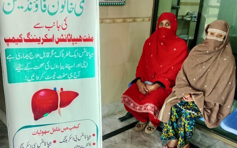 Two women waiting beside a banner for the Salma Khatoon Foundation hepatitis screening camp at the health center in Sahiwal